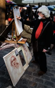 Cette photographie montre un vieil homme, sujet principal, tenant une planche à dessin et observant ses œuvres. La composition met en avant ses peintures posées au sol et sur des chevalets. Le cadrage est serré, centré sur lui et ses tableaux.La lumière semble naturelle, légèrement tamisée, créant des ombres douces. Les couleurs dominantes sont sobres : tons beiges, gris et rouges, avec des touches de noir et blanc dans les dessins.L’ambiance est intimiste et artistique, évoquant un marché ou une rue animée, avec une touche de mélancolie et de créativité.
