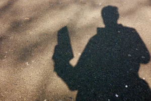 Cette photographie capture une silhouette humaine projetée sur une surface enneigée ou givrée. Le sujet principal est cette ombre, tenant un objet ressemblant à un téléphone ou une caméra. La composition est épurée, centrée sur cette ombre contrastée.Le cadrage est serré, mettant en valeur les contours nets du sujet. La lumière semble naturelle et rasante, créant un fort contraste entre ombre et fond. Les couleurs dominantes sont le noir (silhouette) et le blanc (surface), avec des nuances grises.Le style est minimaliste et épuré, évoquant une ambiance froide et contemplative.