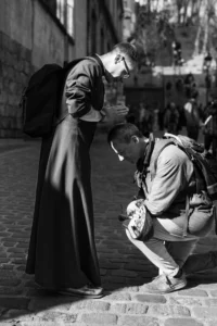Cette photographie en noir et blanc met en scène deux personnes comme sujets principaux : un homme en robe (probablement un moine) et un autre agenouillé, qui semble lui tendre un objet.La composition est dynamique, avec un cadrage serré sur les deux personnages, isolés du reste de la scène. La lumière naturelle, douce mais contrastée, accentue les textures et les ombres, créant une ambiance solennelle et presque théâtrale. Les tons dominants varient du noir profond au gris clair, renforçant le côté dramatique.Le style rappelle le photojournalisme documentaire, évoquant une scène authentique et chargée d’émotion.