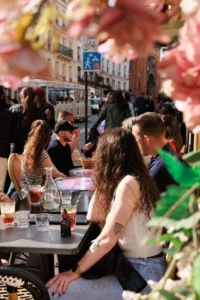 Cette photographie capture un groupe de personnes en extérieur, assis autour de tables dans un café animé. Le sujet principal semble être une femme avec des cheveux longs, tournée vers son compagnon.La composition est dynamique, avec un cadrage serré sur les personnes en premier plan et un arrière-plan flou mais visible. La lumière est naturelle et douce, typique d’une journée ensoleillée. Les couleurs dominantes sont des tons chauds et naturels : verts, bruns, et teintes pastel des vêtements.Le style est documentaire et spontané, évoquant une ambiance conviviale et détendue, presque estivale.