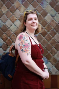 Cette photographie met en scène une personne comme sujet principal, souriante, avec un tatouage floral sur le bras. Elle porte une robe marron et un sac à dos.La composition est simple et centrée, avec un cadrage serré sur le haut du corps. L’arrière-plan texturé et coloré en losanges ajoute un contraste visuel.La lumière est naturelle et douce, éclairant le sujet de manière homogène. Les couleurs dominantes sont les tons chauds (marron, rouge) et les teintes pastel du tatouage.Le style est naturel et décontracté, créant une ambiance conviviale et chaleureuse.