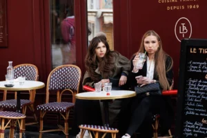 Cette photographie montre deux femmes assises à une terrasse de café. Le sujet principal est leur interaction, captée de manière naturelle.La composition est équilibrée, avec un cadrage serré sur les deux femmes et leur environnement immédiat. Les tables, chaises et menu en arrière-plan ajoutent de la profondeur.La lumière est douce et naturelle, probablement en journée, créant des ombres légères. Les couleurs dominantes sont les tons neutres (beige, gris) avec des touches de rouge et noir, renforçant une ambiance urbaine et élégante.Le style est réaliste et documentaire, évoquant une scène du quotidien. L’ambiance est calme et intimiste.