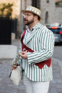 Cette photographie met en scène un homme comme sujet principal, vêtu d’un costume à rayures vertes et blanches, une cravate rouge et un chapeau de paille. Il tient un sac en osier et un accessoire en forme de bouteille.La composition est simple et centrée, avec un cadrage serré sur le sujet. La lumière semble naturelle, légèrement tamisée, créant des ombres douces. Les couleurs dominantes sont les verts, blancs et rouges, apportant une touche vintage et festive.Le style évoque un mélange de décontracté et de théâtral, tandis que l’ambiance suggère une touche humoristique ou décalée, peut-être inspirée d’un personnage de fiction ou d’un événement particulier.