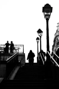 Cette photographie en noir et blanc capture des silhouettes sur un escalier métallique urbain. Le sujet principal est un groupe de personnes descendant ou montant les marches, avec une personne assise en bas à droite.La composition repose sur des lignes verticales (lampadaires) et horizontales (escalier, garde-corps), créant une structure géométrique. Le cadrage est serré, mettant l’accent sur les contrastes d’ombres et de lumières.La lumière, douce et diffuse, accentue les contours et ajoute une ambiance mélancolique. Les tons dominants sont les noirs profonds et les gris, renforçant le côté dramatique et intemporel. Le style est épuré et cinématographique.