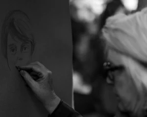 Cette photographie montre un artiste en train de dessiner un portrait au fusain ou au crayon. Le sujet principal est le processus créatif, avec une main tenant le crayon et le visage esquissé en cours de réalisation.La composition juxtapose deux plans : le dessin en cours à gauche et le profil flou de l’artiste à droite. Le cadrage est serré sur les mains et le visage dessiné.La lumière est douce et contrastée, créant des ombres marquées. Les couleurs dominantes sont les tons de gris, typiques d’une image en noir et blanc. L’ambiance est intime et studieuse, évoquant concentration et artisanat.