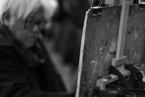 Cette photographie en noir et blanc met en scène un sujet flou en arrière-plan, probablement une personne âgée. Le premier plan montre un objet métallique rouillé, peut-être une structure industrielle ou un meuble abîmé, avec des détails texturés et des éléments de fixation.Le cadrage est serré, centré sur les textures et les formes géométriques. La lumière est douce mais contrastée, créant des ombres marquées et accentuant les reliefs. L’ambiance est mélancolique et nostalgique, renforcée par les tons de gris et les dégradés. Le style rappelle le photojournalisme documentaire ou une approche artistique introspective.
