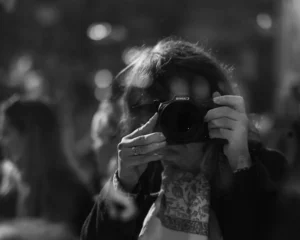 Cette photographie en noir et blanc met en scène une personne comme sujet principal, prise en train de prendre une photo avec un appareil compact. Le cadrage est serré sur ses mains et son visage partiellement caché, créant une ambiance intime et mystérieuse. L’arrière-plan flou suggère une foule, ajoutant une dimension sociale et dynamique.La lumière semble naturelle, avec des contrastes marqués entre ombres et clartés. Le style est documentaire et spontané, renforcé par le flou artistique. Les tons dominants, en niveaux de gris, accentuent les textures et les émotions. L’ambiance est à la fois urbaine et intime, évoquant une scène du quotidien.