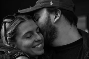 Cette photographie met en scène un couple en train de s'embrasser tendrement. Le sujet principal est leur complicité et leur affection.La composition est simple et centrée, avec un cadrage serré sur leurs visages. La lumière douce et naturelle accentue les traits et crée des contrastes subtils. En noir et blanc, les tons dominants varient du clair au foncé, mettant en valeur les textures et les émotions.Le style est intime et spontané, évoquant une ambiance chaleureuse et romantique.