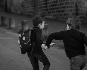 Cette photographie en noir et blanc met en scène deux enfants comme sujets principaux. L’un, avec un sac à dos, semble courir ou jouer, tandis que l’autre tend la main vers lui.La composition est dynamique, centrée sur les enfants en mouvement. Le cadrage serré isole les sujets, avec un arrière-plan flou de bâtiments et de palettes.La lumière est naturelle, douce mais contrastée, accentuant les ombres et les formes. Les tons dominants sont les gris et noirs, typiques du noir et blanc, renforçant une ambiance à la fois réaliste et intemporelle. Le style est documentaire, captant un instant spontané et authentique.