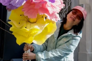 Cette photographie met en scène une personne tenant un ballon en forme de fleur géante, jaune et rose. Le sujet principal est centré, avec un cadrage serré qui isole la personne et son accessoire.La lumière semble naturelle, douce et légèrement tamisée. Les couleurs dominantes sont vives : jaune, rose et des touches de vert (vêtement). Le style est décontracté et joyeux, évoquant une ambiance festive ou ludique, peut-être liée à une célébration ou un événement créatif.