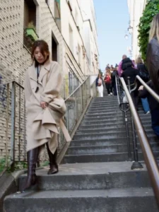Cette photographie montre une femme comme sujet principal, descendant un escalier extérieur dans une tenue élégante : un long manteau beige et des bottes. La composition met en valeur les lignes verticales des bâtiments et des marches, encadrant la scène. Le cadrage est serré, centré sur la femme, avec une profondeur de champ qui floute légèrement l’arrière-plan.La lumière semble naturelle, probablement en journée, avec des ombres douces. Les couleurs dominantes sont les tons neutres (beige, gris) et quelques touches de vert et bleu. Le style est sobre et urbain, évoquant une ambiance moderne et élégante, presque cinématographique.
