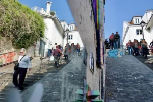 Cette photographie montre une rue pentue et étroite dans un quartier résidentiel. Le sujet principal semble être les passants et l’architecture locale.La composition juxtapose deux images quasi identiques, révélant un effet de miroir ou de superposition. Le cadrage est serré sur les bâtiments blancs et les escaliers pavés.La lumière est naturelle et claire, suggérant un jour ensoleillé. Les couleurs dominantes sont les tons blancs des murs, les gris des pavés et les touches de vert des plantes.Le style est documentaire, avec une ambiance vivante et quotidienne, reflétant une scène de vie urbaine paisible.