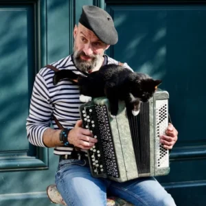 Cette photographie met en scène un homme comme sujet principal, assis et jouant de l'accordéon. Un chat noir est posé sur l’instrument, ajoutant une touche de spontanéité.La composition est simple et naturelle, avec un cadrage serré qui isole les deux sujets. La lumière semble douce et naturelle, éclairant le visage et l’instrument sans créer d’ombres marquées.Les couleurs dominantes sont sobres : bleu et gris pour les vêtements, noir pour le chat, et des tons neutres pour l’accordéon. Le style est documentaire et chaleureux, évoquant une ambiance intimiste et apaisante.