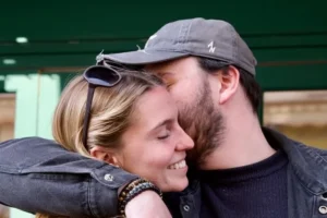 Cette photographie capture un couple en train de s'embrasser tendrement. Le sujet principal est centré sur leurs visages, avec un cadrage serré et une composition intime.La lumière semble naturelle, douce et légèrement tamisée, créant une ambiance chaleureuse. Les couleurs dominantes sont les tons neutres (gris du pull et du chapeau) et quelques touches de vert en arrière-plan.Le style est simple et spontané, évoquant une scène authentique et émotionnelle. L'ambiance est romantique et affectueuse, renforcée par le contact physique et les expressions des visages.