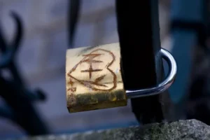 Cette photographie met en scène un ** cadenas personnalisé** attaché à une structure métallique, probablement un pont ou une rambarde. Le cadenas porte une gravure stylisée d’un cœur et d’un motif rappelant une clé ou un symbole abstrait.La **composition** est simple et centrée sur le cadenas, avec un **cadrage serré**. La lumière naturelle, douce et légèrement tamisée, crée des contrastes subtils. Les **couleurs dominantes** sont des tons chauds (or, brun) et des teintes froides (gris métallique, bleu de l’arrière-plan flou).Le style est **minimaliste et poétique**, évoquant une ambiance **romantique et mélancolique**, typique des cadenas d’amour.