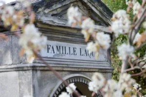 Cette photographie met en scène un mausolée ou une tombe portant l'inscription **« FAMILLE LAGU... »**. Le sujet principal est ce monument funéraire de style classique, avec des lignes épurées et une architecture sobre.Le cadrage est serré, centré sur l'inscription, avec des branches fleuries en premier plan, légèrement floues. La lumière naturelle, douce et tamisée, crée une ambiance paisible et mélancolique. Les couleurs dominantes sont les tons gris-beige du monument, contrastés par le blanc des fleurs. L'ensemble évoque une atmosphère solennelle et intemporelle.