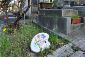 Cette photographie met en scène un **pinceau en céramique peint** posé sur un sol en pierre, entouré de végétation et de fleurs jaunes. Le sujet principal est ce pinceau décoratif, évoquant l’art.La **composition** est simple et naturelle, avec un cadrage serré sur l’objet, entourée d’éléments organiques. La lumière semble douce et naturelle, créant des ombres légères. Les **couleurs dominantes** sont les tons pastel du pinceau, le vert de la végétation et le jaune des fleurs.L’ambiance est **poétique et mélancolique**, mêlant créativité et nature, dans un style presque documentaire.