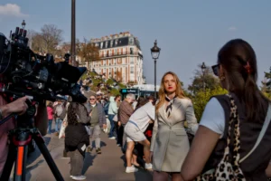 Cette photographie montre une femme en costume gris comme sujet principal, discutant avec une autre personne. La composition inclut plusieurs individus en arrière-plan, suggérant une scène publique ou un événement médiatique.Le cadrage est serré sur les deux femmes, avec une caméra de télévision visible à gauche. La lumière est naturelle et claire, typique d’un jour ensoleillé. Les couleurs dominantes sont des tons neutres (gris, beige) avec des touches de vert et de bleu dans l’arrière-plan.L’ambiance semble professionnelle et dynamique, évoquant une interview ou une couverture médiatique en extérieur.