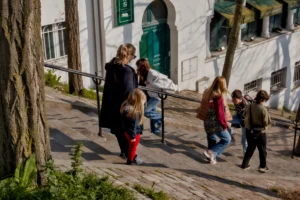 Cette photographie capture un groupe de personnes, principalement des enfants et des adultes, marchant sur une allée en pente pavée. Le sujet principal semble être les interactions entre ces individus.La composition est dynamique, avec un cadrage légèrement bas, mettant en valeur les visages et les mouvements. La lumière naturelle, douce et légèrement dorée, crée des ombres nettes. Les couleurs dominantes sont les tons neutres (beige, gris) avec des éclats de rouge et vert.Le style est documentaire et spontané, évoquant une ambiance chaleureuse et vivante dans un cadre urbain ou semi-urbain.