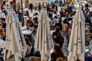 Cette photographie montre une scène animée de terrasse de café en extérieur. Le sujet principal est une série de parasols blancs, marqués du logo "WHITE HUT". La composition est dynamique, avec une foule de personnes assises et debout, créant une ambiance sociale et conviviale. Le cadrage est légèrement en plongée, offrant une vue d'ensemble.La lumière semble naturelle, probablement en journée. Les couleurs dominantes sont les tons neutres (blanc, beige) et les couleurs chaudes des vêtements des gens. Le style est documentaire et réaliste, capturant un moment de vie quotidienne. L’ambiance est chaleureuse et animée.