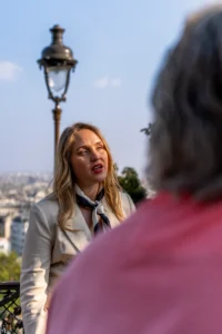Cette photographie met en scène une femme comme sujet principal, vêtue d’un blazer blanc et d’une cravate, en train de parler avec enthousiasme. Le cadrage est serré sur elle et une partie d’un homme en arrière-plan, vêtu de rose.La composition joue sur les contrastes entre les visages et l’arrière-plan urbain flou. La lumière est naturelle, douce et légèrement latérale, éclairant les sujets sans ombres marquées. Les couleurs dominantes sont les tons pastel (blanc, rose) et les teintes neutres de la ville en arrière-plan.Le style est réaliste et spontané, évoquant une ambiance chaleureuse et conversationnelle, presque cinématographique.