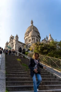 Cette photographie montre une femme assise en haut d’un escalier monumental menant au Sacré-Cœur de Paris. Le sujet principal est elle, vêtue d’un jean et d’un pull, avec un sac à dos. La composition met en valeur les marches en pierre et l’architecture du bâtiment en arrière-plan.Le cadrage est serré sur la femme, tout en intégrant l’édifice emblématique. La lumière est naturelle et douce, créant des ombres légères. Les couleurs dominantes sont les tons beiges des pierres, les bleus du ciel et les verts des arbres. L’ambiance est paisible et touristique, avec une touche de sérénité.