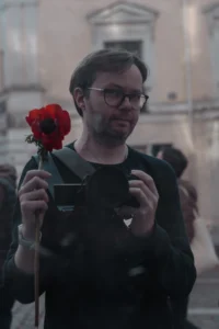 Cette photographie montre un homme comme sujet principal, tenant une rose rouge à la main. Le cadrage est serré, centré sur son visage et ses mains, avec un arrière-plan flou où l’on devine des bâtiments et des personnes.La lumière semble naturelle, tamisée, créant des contrastes doux. Les couleurs dominantes sont les tons sombres de ses vêtements, contrastant avec le rouge vif de la rose. Le style est sobre et réaliste, évoquant une ambiance intime et légèrement mélancolique, peut-être liée à une manifestation ou un événement solennel.