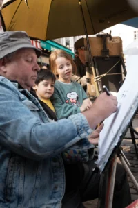 Cette photographie montre un homme aidant deux enfants à dessiner sur une feuille posée sur une table. Le sujet principal est cette interaction intergénérationnelle.La composition est serrée, centrée sur les mains et les visages. Le cadrage est rapproché, mettant en valeur les détails et les expressions. La lumière semble naturelle et douce, éclairant les sujets de manière homogène.Les couleurs dominantes sont des tons neutres (bleu du vêtement de l’homme, gris clair) avec des touches de couleurs vives sur les vêtements des enfants. L’ambiance est chaleureuse et conviviale, évoquant un moment d’apprentissage et de partage.