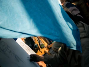 Cette photographie montre une personne en train de dessiner sous une bâche bleue. Le sujet principal est le bras et la main tenant un stylo, en action sur une feuille posée sur une surface.La composition est serrée, centrée sur le geste précis. Le cadrage est rapproché, mettant en valeur les détails. La lumière semble naturelle, avec des contrastes doux. Les couleurs dominantes sont les teintes bleutées de la bâche, les tons neutres de la feuille et les couleurs vives du vêtement jaune-vert.Le style est documentaire et réaliste, capturant un moment spontané. L’ambiance est calme et concentrée.
