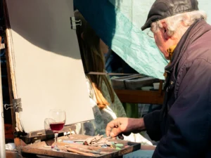 Cette photographie montre un homme âgé, sujet principal, en train de peindre ou de travailler sur une petite toile ou un objet. Le cadrage est serré, mettant en valeur ses mains et son activité.La lumière naturelle éclaire la scène, créant des contrastes doux. Les couleurs dominantes sont neutres (beige, gris) avec des touches de rouge (vin) et de vert (feuilles, palette).L’ambiance est intimiste et artisanale, évoquant le travail méticuleux et créatif. Le style semble documentaire, capturant un moment authentique de création.