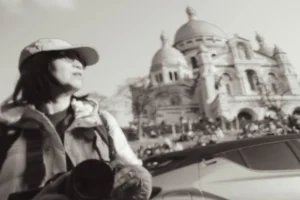Cette photographie en noir et blanc met en scène une femme comme sujet principal, souriante et détendue, vêtue d’un chapeau et d’un manteau, assise sur un véhicule.La composition montre en arrière-plan la basilique du Sacré-Cœur de Paris, partiellement visible. Le cadrage est serré sur la femme, avec une profondeur de champ qui floute légèrement le fond.La lumière est douce, naturelle, créant des contrastes modérés. Les tons dominants sont les gris et noirs, typiques du noir et blanc, renforçant une ambiance nostalgique et poétique. Le style est documentaire et spontané, évoquant une atmosphère paisible et touristique.