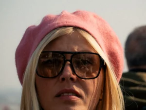 Cette photographie met en scène une femme comme sujet principal, vêtue d’un bonnet rose et portant des lunettes de soleil. Le cadrage est serré, centré sur son visage, avec une légère floueur en arrière-plan.La lumière semble naturelle, bien que contrastée, créant des ombres douces. Les couleurs dominantes sont les tons chauds (rose du bonnet, blond des cheveux) et les teintes neutres (peau, lunettes).Le style est simple et direct, évoquant un moment du quotidien. L’ambiance est à la fois décontractée et légèrement mystérieuse grâce aux lunettes et à l’expression neutre.