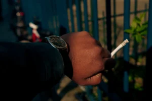 Cette photographie met en scène une main tenant une cigarette allumée, avec une montre visible au poignet. Le sujet principal est donc cette main, partiellement vêtue d’un pull noir.Le cadrage est serré, centré sur la main et l’objet, avec un arrière-plan flou montrant une barrière et des éléments urbains. La lumière est tamisée, créant des contrastes doux et une ambiance nocturne ou peu éclairée. Les couleurs dominantes sont les teintes sombres (noir, gris) avec des touches de bleu et de vert en arrière-plan.Le style est réaliste et sobre, évoquant une ambiance contemplative et intime.