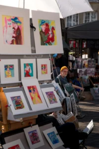 Cette photographie montre une personne assise derrière un étalage d’art. Le sujet principal est l’artiste ou vendeur, entouré de ses œuvres colorées.La composition met en avant des tableaux abstraits et colorés, disposés en plusieurs rangées. Le cadrage est serré sur l’étal et l’artiste, avec une légère profondeur de champ.La lumière semble naturelle, mettant en valeur les couleurs vives des toiles. Les couleurs dominantes sont vives et contrastées : jaunes, rouges, bleus et verts.L’ambiance est artistique et chaleureuse, évoquant un marché ou une foire d’art. Le style est spontané et vivant.