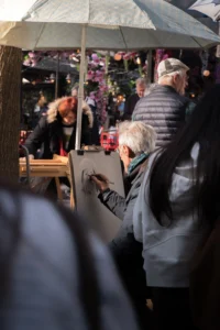 Cette photographie capture une scène de marché ou de fête nocturne. Le sujet principal est un groupe de personnes âgées sous un parapluie transparent, observant des décorations ou des stands illuminés.La composition montre un cadrage serré, mettant en avant les visages et les détails des vêtements. La lumière est douce et tamisée, créant une ambiance chaleureuse et festive. Les couleurs dominantes sont les tons froids (gris, bleu) contrastés avec des éclats chauds (rouges, oranges) des décorations en arrière-plan.Le style est documentaire et naturel, évoquant une ambiance conviviale et intimiste.