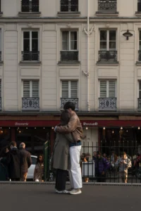 Cette photographie capture un couple s'embrassant devant un immeuble parisien. Le sujet principal est central, mis en valeur par un cadrage serré et une composition en profondeur.La lumière est douce et naturelle, typique d'une journée urbaine. Les couleurs dominantes sont neutres : gris, beige et blanc, avec des touches de rouge et de noir pour les enseignes et détails architecturaux. Le style est réaliste et documentaire, reflétant une ambiance intimiste et romantique dans un cadre urbain animé.