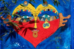 Cette photographie met en scène un cœur coloré et stylisé, sujet principal, peint sur un support mural bleu. Le cœur est décoré de motifs variés : yeux géométriques, mains et éléments graphiques colorés. La composition est symétrique, avec des éléments artistiques et symboliques.Le cadrage est serré, centré sur le cœur. La lumière est naturelle, mettant en valeur les couleurs vives et les détails. Les couleurs dominantes sont le bleu, le rouge, le jaune et le vert, créant un style artistique et joyeux. L’ambiance est festive, positive et pleine d’énergie.