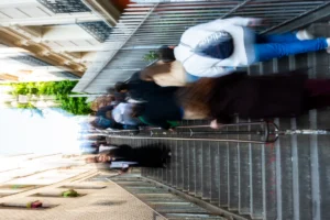 Cette photographie capture un **marché animé** avec pour **sujet principal** un vendeur en mouvement, flou, tenant des objets (probablement des fruits ou légumes).La **composition** est dynamique, avec une perspective basse et un cadrage serré sur le comptoir métallique. La **lumière** est naturelle, légèrement contrastée, créant des ombres douces. Les **couleurs dominantes** sont des tons verts, gris et blancs, avec des éclats de bleu et de blanc (emballages).Le **style** est réaliste et spontané, évoquant une **ambiance** vivante et quotidienne, typique d’un marché local.