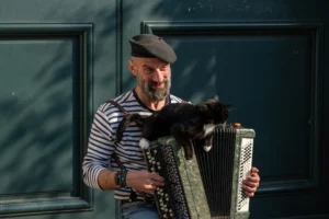 Cette photographie met en scène un homme âgé, sujet principal, jouant de l'accordéon tout en tenant un chat noir. La composition est simple et centrée, avec un cadrage serré sur le visage et les mains de l'homme.La lumière naturelle, douce et légèrement latérale, éclaire son visage, créant des ombres douces. Les couleurs dominantes sont sobres : bleu et blanc pour le vêtement strié de l'homme, noir pour le chat et l'accordéon, avec des touches métalliques grises.Le style est réaliste et spontané, évoquant une ambiance chaleureuse et intimiste, presque poétique.