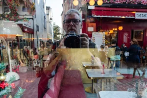 Cette photographie met en scène un homme vêtu de rouge et or, tenant un objet doré, probablement une offrande. Le sujet principal semble être lié à une cérémonie religieuse ou culturelle.La composition est serrée, centrée sur son visage et ses mains. Le cadrage est rapproché, avec un arrière-plan flou montrant des éléments colorés et des gens en mouvement. La lumière est naturelle et semble douce, bien que légèrement contrastée.Les couleurs dominantes sont le rouge, l’or et le blanc, créant une ambiance riche et festive. Le style est documentaire, capturant un moment authentique et spontané dans une ambiance vibrante et traditionnelle.