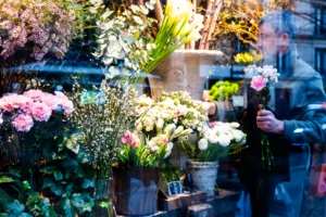 Cette photographie met en scène un **marché de fleurs**. Le **sujet principal** est une variété de bouquets et de plantes disposés dans des pots et des paniers. La **composition** est dynamique, avec des éléments superposés et un léger flou d’arrière-plan.Le **cadrage** est serré, centré sur les fleurs, tandis que la **lumière** naturelle, douce, met en valeur les teintes. Les **couleurs dominantes** sont des tons pastel (rose, blanc, vert) et quelques touches vives.Le **style** est réaliste et spontané, évoquant une ambiance **vivante et chaleureuse**, typique d’un marché artisanal.