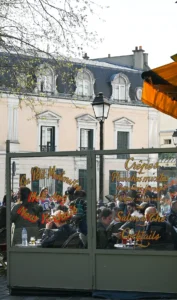 Cette photographie capture une scène de terrasse de café en extérieur. Le sujet principal est un groupe de personnes assises sous des parasols orange, discutant et profitant de leur boisson.La composition montre une vue en profondeur, avec un cadrage serré sur les clients et un arrière-plan de bâtiments classiques. La lumière est naturelle et douce, typique d'une journée ensoleillée. Les couleurs dominantes sont les tons chauds (orange des parasols, beige des murs) et les verts des plantes grimpantes.Le style est réaliste et spontané, évoquant une ambiance conviviale et détendue, caractéristique des cafés urbains.