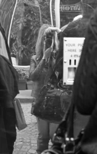 Cette photographie en noir et blanc capture une scène urbaine floue. Le sujet principal semble être une personne avec une longue chevelure blonde, partiellement visible, tenant un sac. La composition est dynamique, avec un cadrage serré et légèrement désordonné. La lumière est tamisée, créant des contrastes doux et des ombres marquées.L’ambiance est intimiste et un peu mystérieuse, renforcée par le flou artistique. Les couleurs dominantes, bien que monochromes, suggèrent des tons gris et noirs. Le style rappelle une approche documentaire ou artistique, avec une touche de spontanéité et de mouvement.