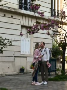 Cette photographie capture un couple marchant dans une rue urbaine. Le sujet principal est ce couple, dont les visages sont floutés, l’un portant un sac à dos et l’autre une veste rose.La composition est simple et naturelle, avec un cadrage serré sur les deux personnes. En arrière-plan, on aperçoit une façade classique et des fleurs en treillis, ajoutant une touche de couleur.La lumière semble douce et naturelle, typique d’une journée ensoleillée. Les couleurs dominantes sont les tons neutres de la pierre et des vêtements, rehaussés par les touches de rose et de vert.L’ambiance est paisible et romantique, évoquant une balade urbaine détendue.