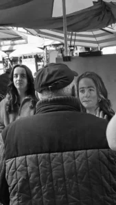 Cette photographie en noir et blanc montre trois personnes en conversation. Le sujet principal semble être les deux femmes en premier plan, dont une porte un béret et un manteau texturé. La composition est serrée, avec un cadrage rapproché mettant l'accent sur les visages et les expressions.La lumière est douce, sans ombres marquées, créant une ambiance naturelle et intime. L'absence de couleurs accentue les contrastes et les textures. Le style est documentaire et spontané, évoquant une scène du quotidien. L'ambiance est chaleureuse et conviviale.
