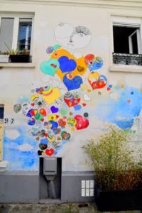 Cette photographie montre un **mural coloré** représentant une **licorne stylisée**, sujet principal. La composition est dynamique, avec des formes organiques et des motifs variés intégrant des visages et des éléments abstraits.Le cadrage est serré, mettant en valeur les détails du mur. La lumière naturelle éclaire uniformément la scène, sans ombre marquée. Les **couleurs dominantes** sont vives : bleus, jaunes, rouges et verts, créant un contraste joyeux.Le style est **artistique et naïf**, évoquant un mélange de pop art et de fantaisie. L’ambiance est **ludique et onirique**, apportant une touche de magie urbaine.