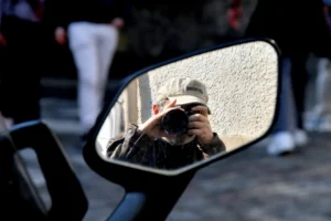 Cette photographie met en scène un **sujet principal** : le reflet d’une personne prenant une photo dans un rétroviseur de scooter. La **composition** est centrée sur ce miroir, créant un effet de symétrie et de profondeur. Le **cadrage** est serré, isolant le rétroviseur et son reflet.La **lumière** semble naturelle, avec des contrastes doux, et une ambiance légèrement grise. Les **couleurs dominantes** sont des tons neutres (gris, noir, beige), avec des touches de blanc et de rouge (vêtements).Le **style** est réaliste et spontané, évoquant une scène urbaine du quotidien. L’**ambiance** est moderne et introspective, mêlant mouvement et immobilité.