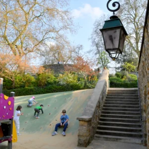 Cette photographie capture un espace vert urbain avec une aire de jeux comme sujet principal. La composition montre une pente herbeuse où des enfants jouent, encadrée par des escaliers en pierre menant vers un lampadaire ancien.Le cadrage inclut une partie des arbres et du ciel, créant une ambiance aérée. La lumière est naturelle, douce et légèrement tamisée, suggérant un moment en fin de journée. Les couleurs dominantes sont les verts de la végétation, les tons gris des pierres et le bleu du ciel, avec des éclats colorés des vêtements des enfants.Le style est documentaire et spontané, évoquant une ambiance détendue et joyeuse.