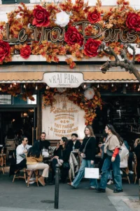 Cette photographie capture l’entrée d’un café parisien nommé "Le Foyer & Paris". Le sujet principal est l’ambiance conviviale des clients assis en terrasse. La composition met en avant l’enseigne lumineuse et les guirlandes florales rouges, encadrant l’espace.Le cadrage est serré sur l’entrée et la terrasse, avec une lumière naturelle douce. Les couleurs dominantes sont les tons chauds (rouge, beige, brun) et les touches de vert et bleu. Le style est réaliste et spontané, évoquant une ambiance chaleureuse et typiquement parisienne, mêlant élégance et simplicité.