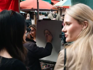 Cette photographie montre deux femmes observant une œuvre d’art en cours de création, probablement un portrait. Le sujet principal est le tableau et les mains de l’artiste qui le dessine.La composition est serrée, centrée sur les visages et le travail artistique. Le cadrage est en plan moyen, mettant en valeur les interactions et les détails.La lumière semble naturelle et douce, éclairant les sujets sans ombres marquées. Les couleurs dominantes sont neutres, avec des touches de rouge et de vert en arrière-plan.L’ambiance est conviviale et artistique, évoquant un événement culturel ou une exposition.